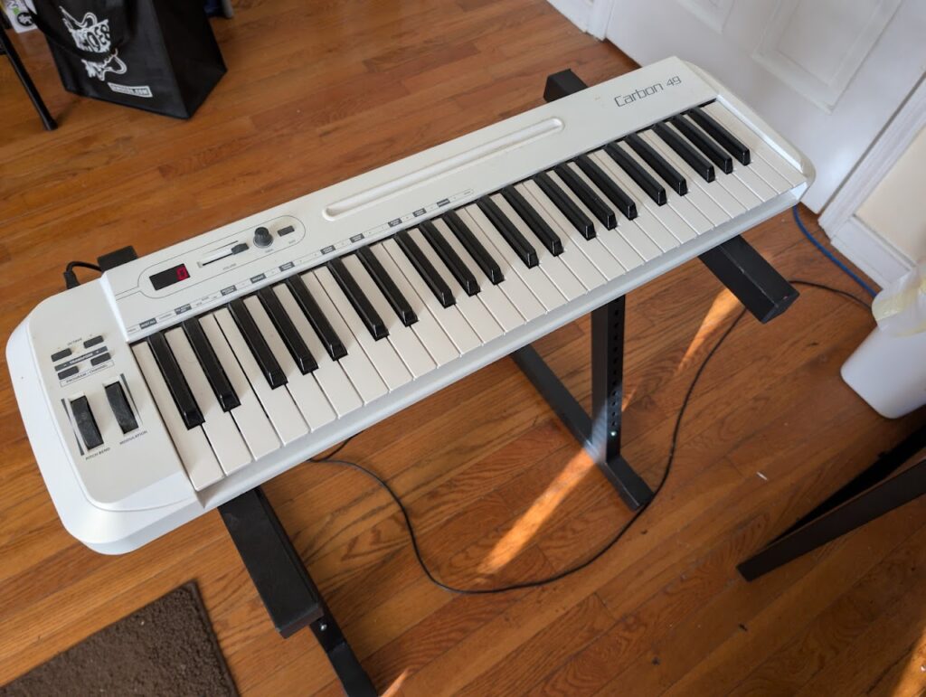 White Samson Carbon 49 musical keyboard on a stand. Picture taken top down with the stand on top of a wooden floor.