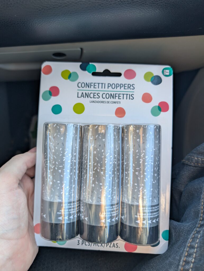 Showing a package of confetti poppers, there are 3 confetti poppers in the package. The text on top of the package says "Confetti Poppers" as well as translation in at least 2 other languages I don't recognize.