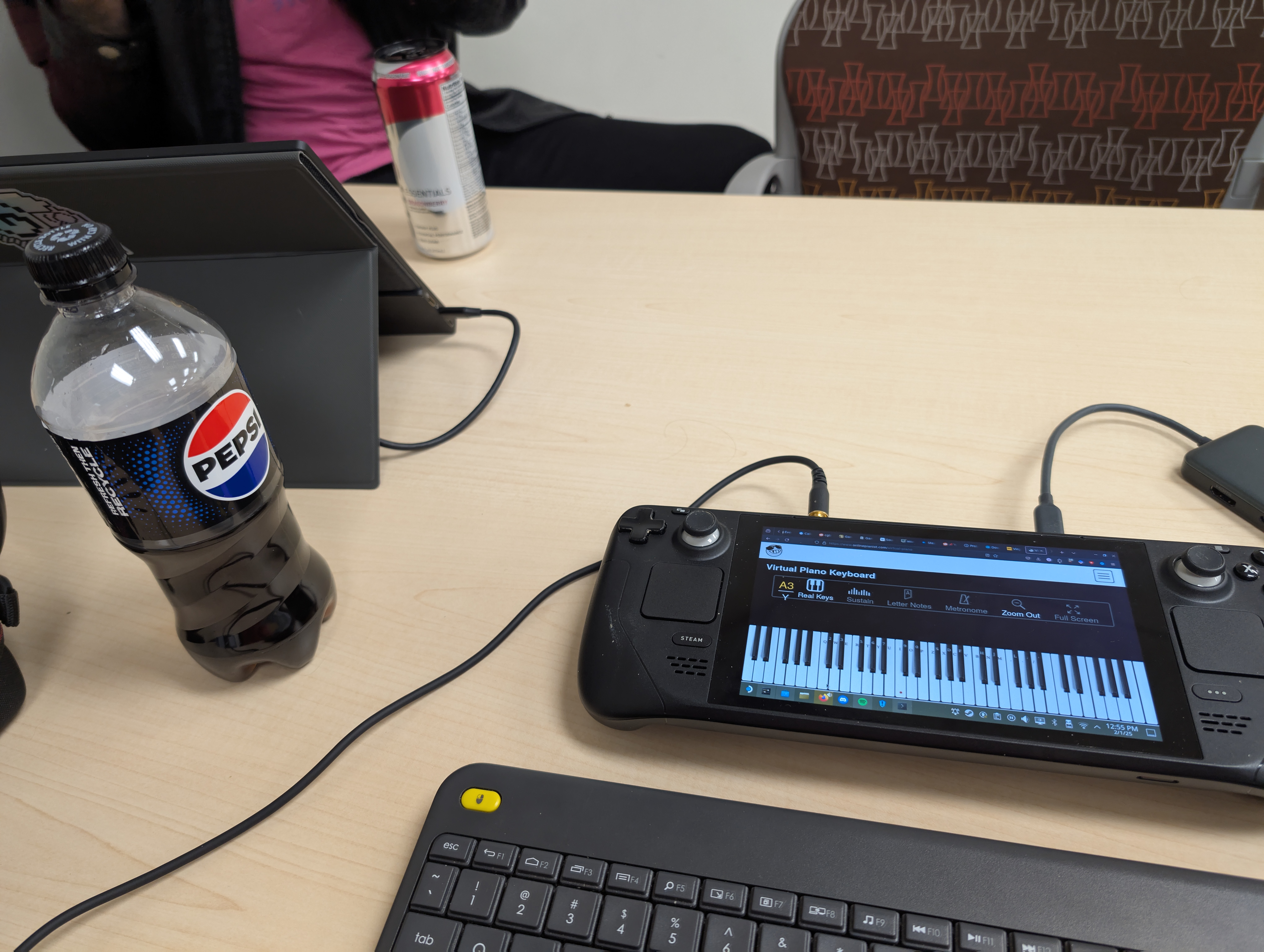 A Woodgrain desk with a laptop towards the left side. In fromt of the lap top is a Pepsi bottle. Towards the right side of the picture on the desk is a steam deck with a piano on the screen. Below the steam deck you partially see a portable computer keyboard. there is a dock and headphone wire connected to the top of the steam deck.