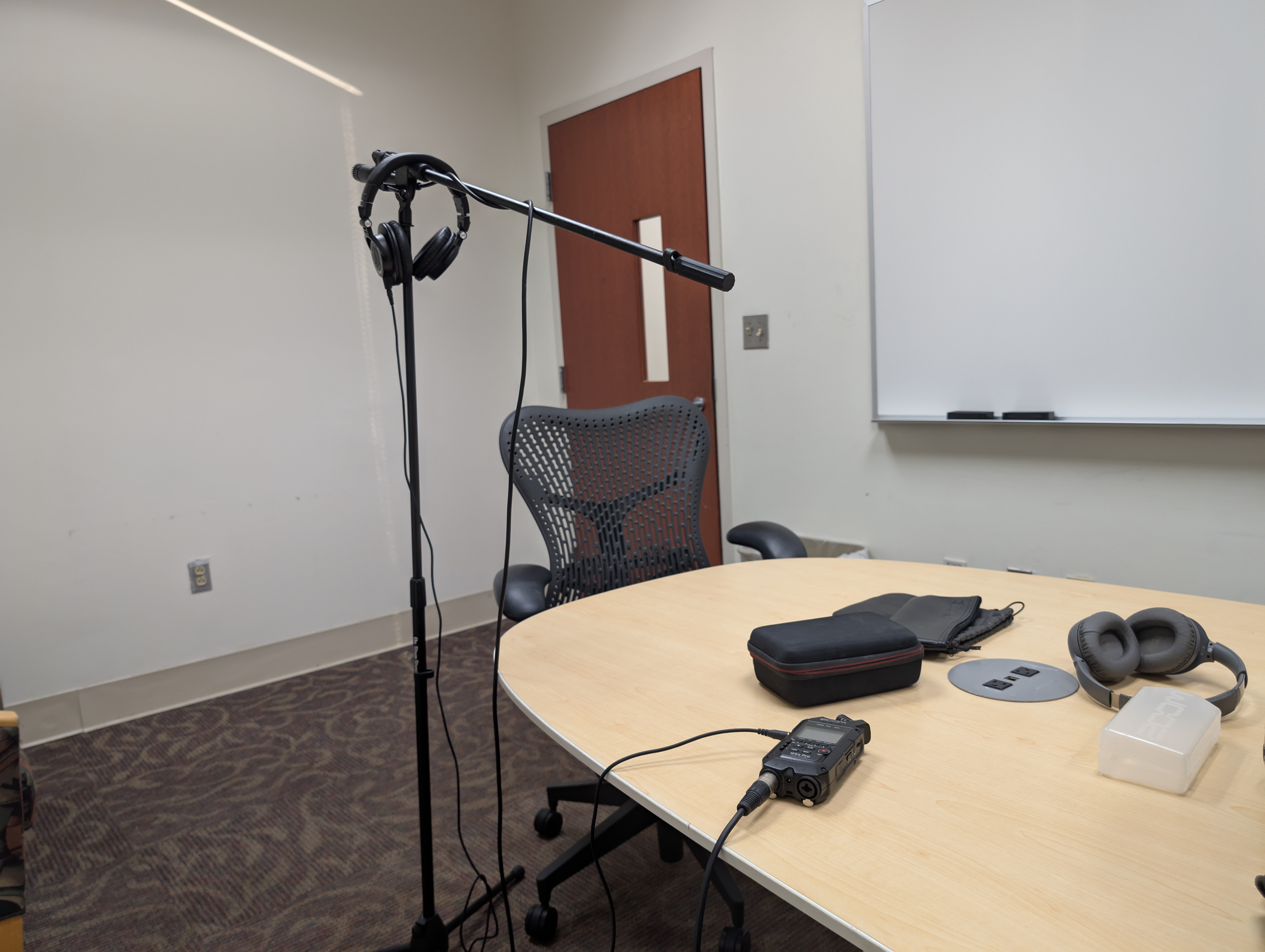 A microphone stand in a mostly empty room. to the right hand side of the mice stand is a woodgrain meeting desk with some headphones, a couple of bags, a hard carrying case, and a zoom H4N audio recorder. PLugged into the audio recorder is headphones and a microphone cable. On the mic stand itself, you see a Shure SM57 microphone and a pair of headphones hanging on the stand.