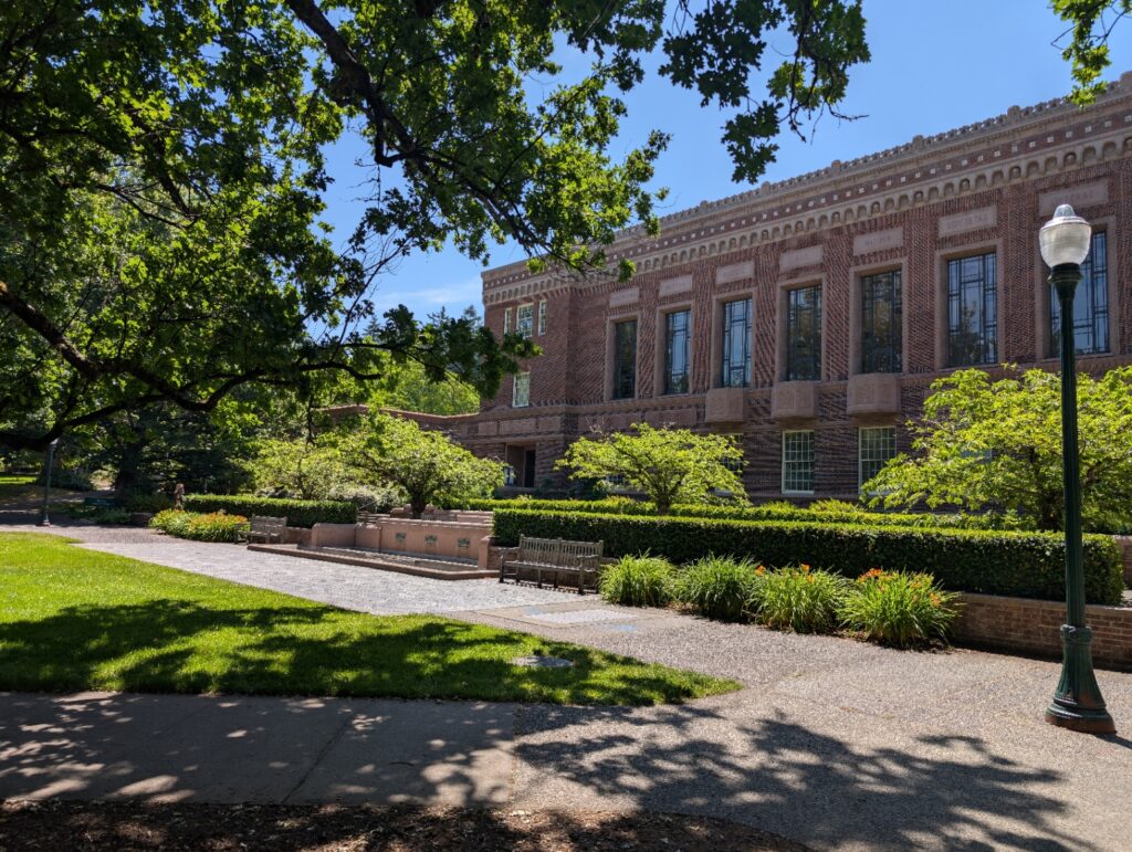 A bright, sunny day in front of Knight library on the University of Oregon campus. You see lots of greenery in front.