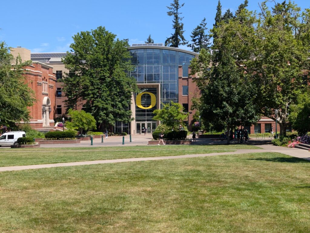 A bright, sunny day in front of Lillis business center on the University of Oregon campus. You see lots of greenery in front. The building displays a big yellow "O".