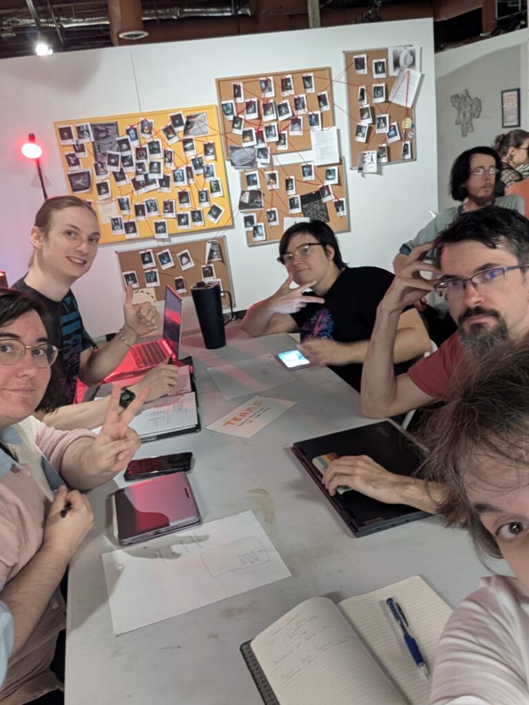 Picture of the entire team gathered around the desk. from left to right, you have Andrew, Tori, myself in the bottom right corner, Galen, and Mickey.

The background is a wall that looks like a crime scene bulletin board from a previous art exhibit.