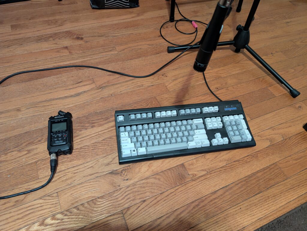 Picture of a standard Keyboard on a wooden floor with a microphone on top of it. They keyboard is black with grey keys. to the left of the keyboard is the zoom H4N audio recorder. 