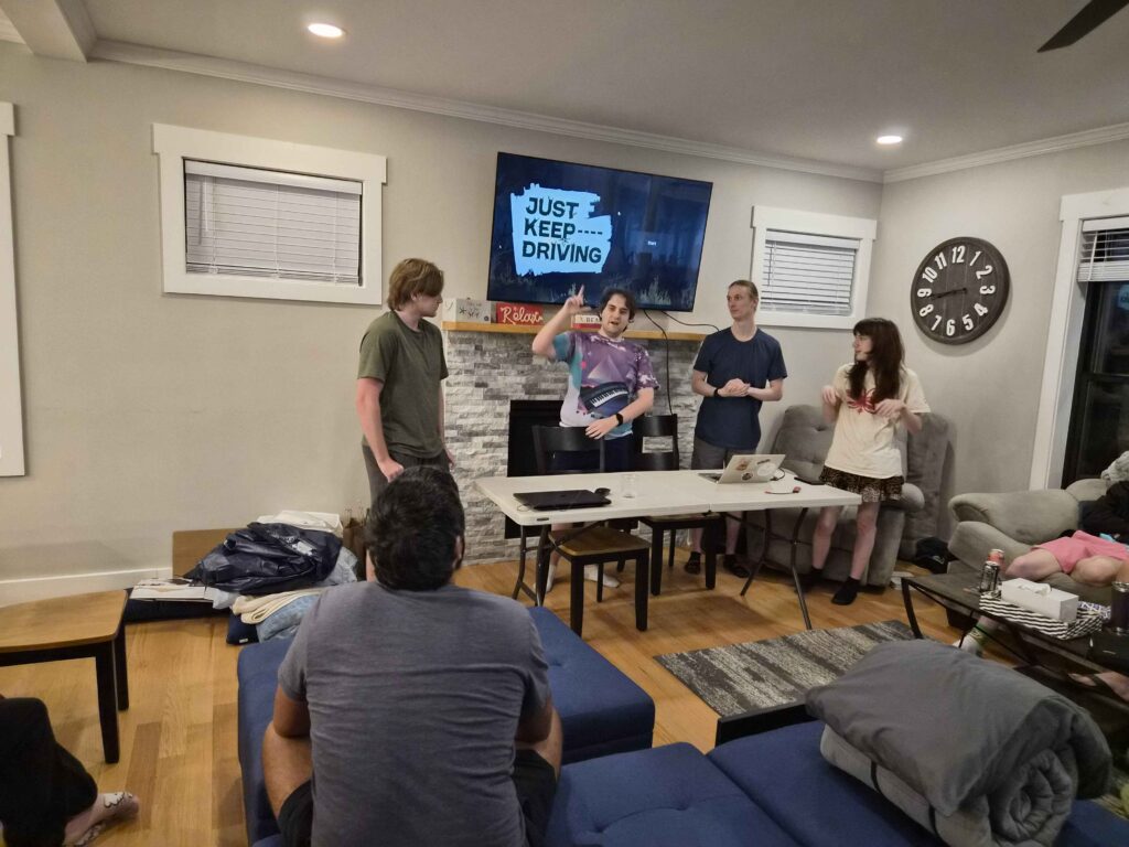 Team presentation. TV above the fireplace showing the logo for "Just Keep Driving" From left to right you have Dylan, Myself, Andrew, and Ella