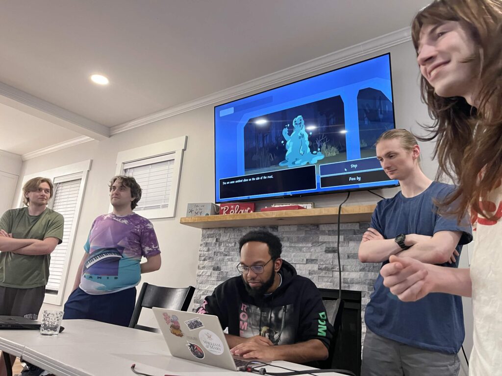 Close up view of the team presentation. Same lineup of people as the previous picture. In the middle of the lineup, you have someone on the laptop play testing the game during the presentation.