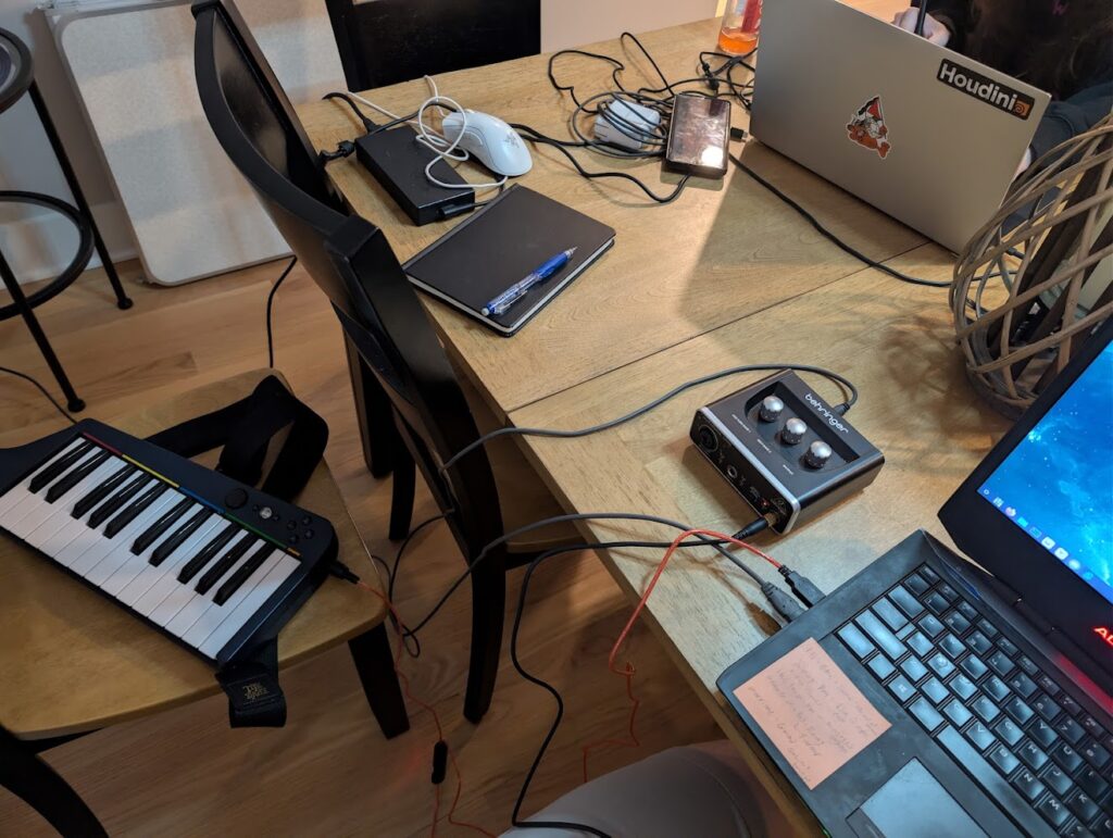 A sprawling mess of a setup. It shows a couple of laptops and cables on a dining room table, with a music keyboard on a nearby chair.